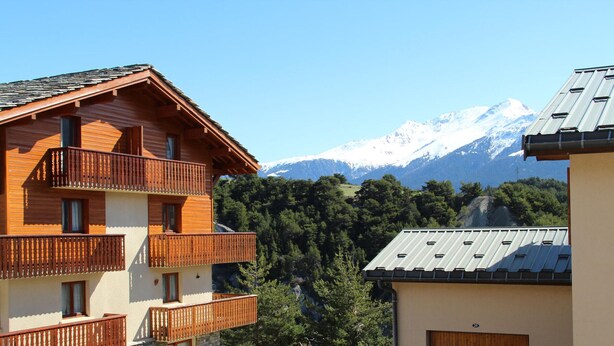 A multi-story wooden building with balconies, a stone foundation, and a view of snow-capped mountains. - Quiet apartment in Aussois, near shuttles and leisure (Aussois)