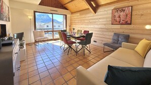 Une pièce chaleureuse avec un plafond en bois, un coin repas avec des chaises, un canapé et une grande fenêtre donnant sur les montagnes.