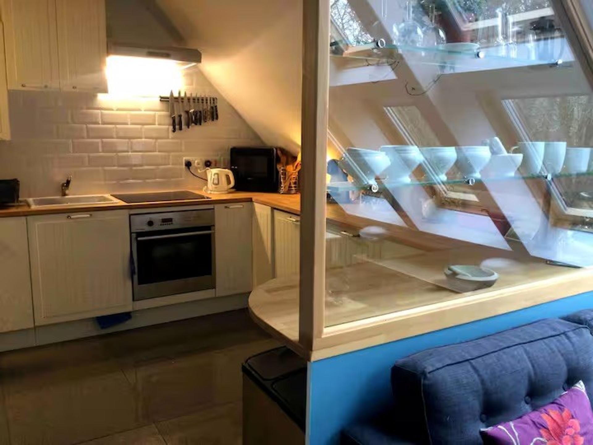 Cozinha moderna com armários brancos, forno embutido e micro-ondas. Há uma janela com vista para o céu e uma parede azul em primeiro plano.
