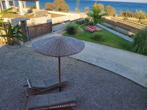 Een terras met een rieten parasol, twee loungesteilen en uitzicht op zee.