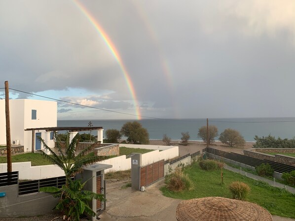 Una zona residencial con vistas al mar, que presenta un edificio blanco con puerta azul, un muro de piedra y un espacio con césped y una cesta tejida.