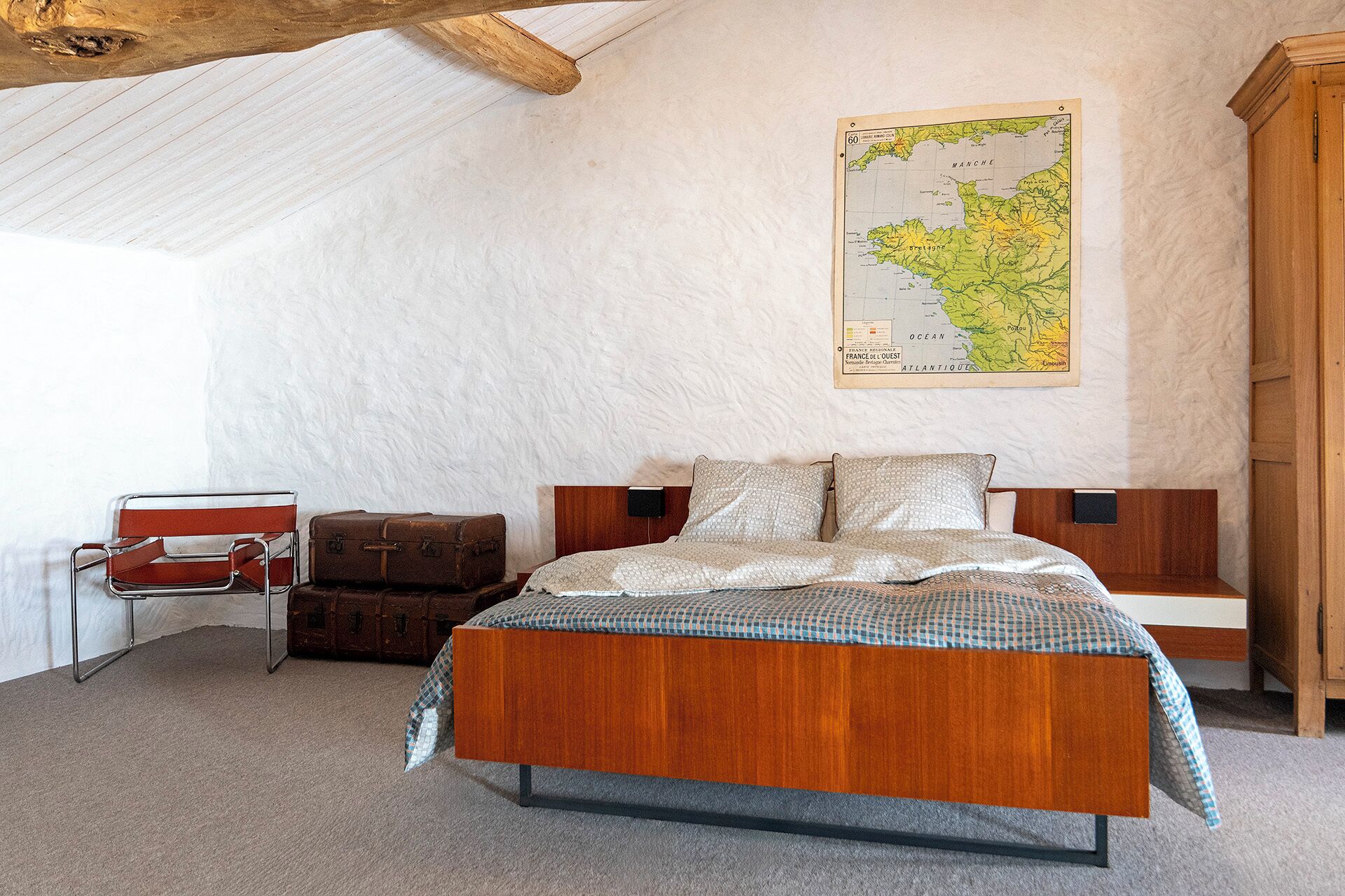 Un dormitorio con cama de madera, un mapa en la pared, una maleta antigua y una silla roja.