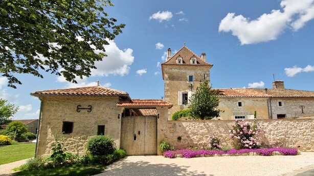 A historic stone building with a tower, a gated entrance, and a well-maintained garden with pink flowers.