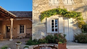 A rustic stone building with a tiled roof, a wooden pergola, and a garden area with potted plants and a fire pit.