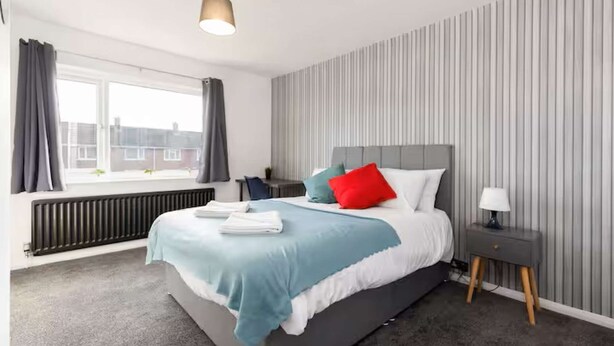 A modern hotel room with a large bed, a bedside table, a radiator, and a window with curtains. - 4 bedroom home in Billingham (Stockton on Tees)