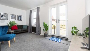 A modern living room with a blue sofa, a grey armchair, and a coffee table. There are two potted plants, a blue and white rug, and a TV stand with a remote control. - 4 bedroom home in Billingham (Stockton on Tees)