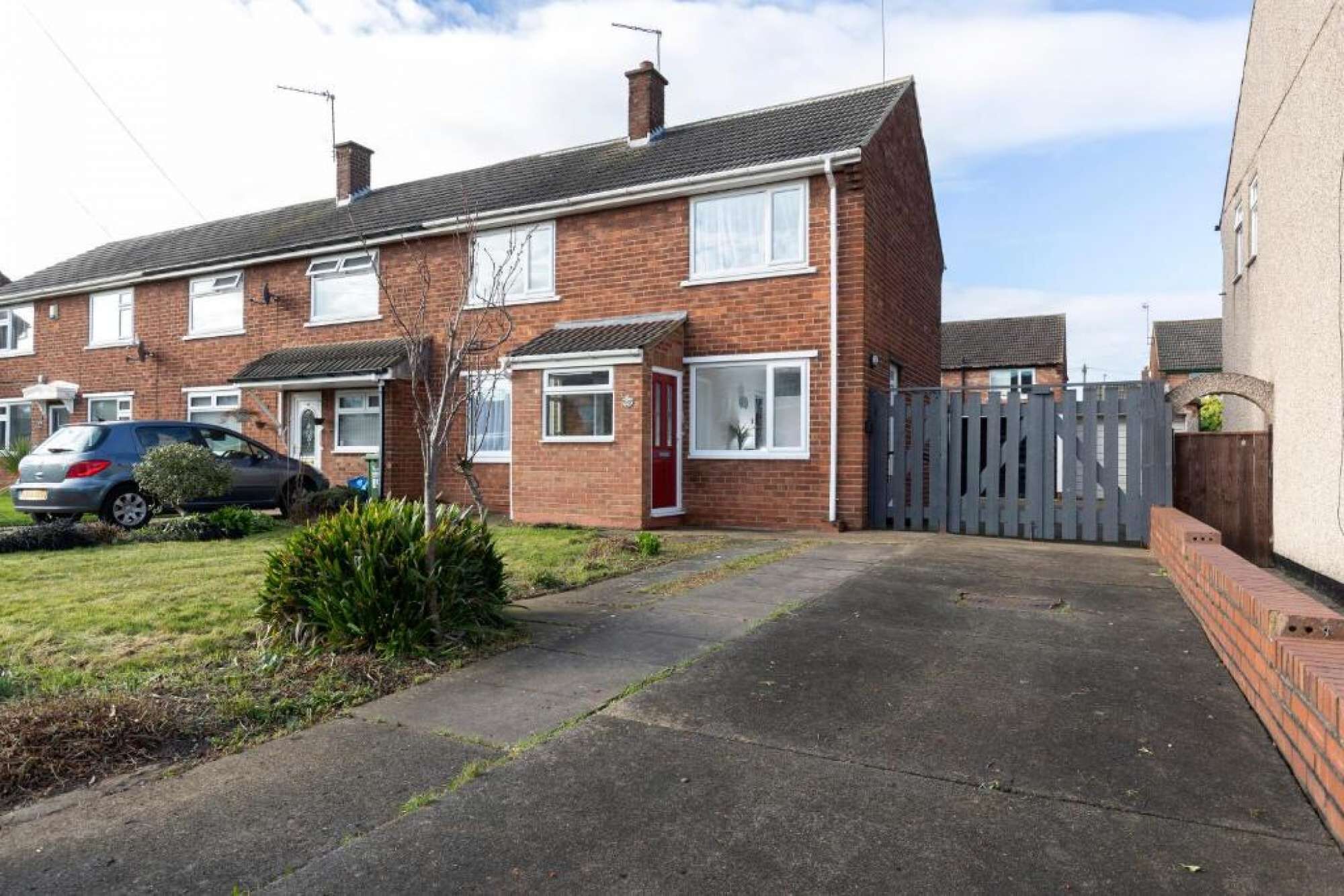 A two-story brick house with a front garden, a car parked in the driveway, and a grey fence.
