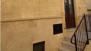 Un bâtiment à la façade en pierre beige, avec une petite fenêtre, une porte dotée d’un panneau de verre et un escalier en métal noir.