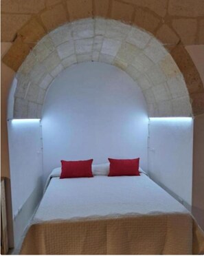 Una habitación con techo abovedado de piedra, una cama con ropa de cama blanca y almohadas rojas, y una pared lisa.