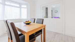 Un comedor con una mesa de madera y cuatro sillas, un cuenco de fruta sobre la mesa y una ventana con cortinas ligeras.