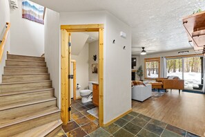 Staircase leads to a bathroom with a toilet and cabinet. The living area features a sofa, coffee table, and large windows offering a view of the outdoors.