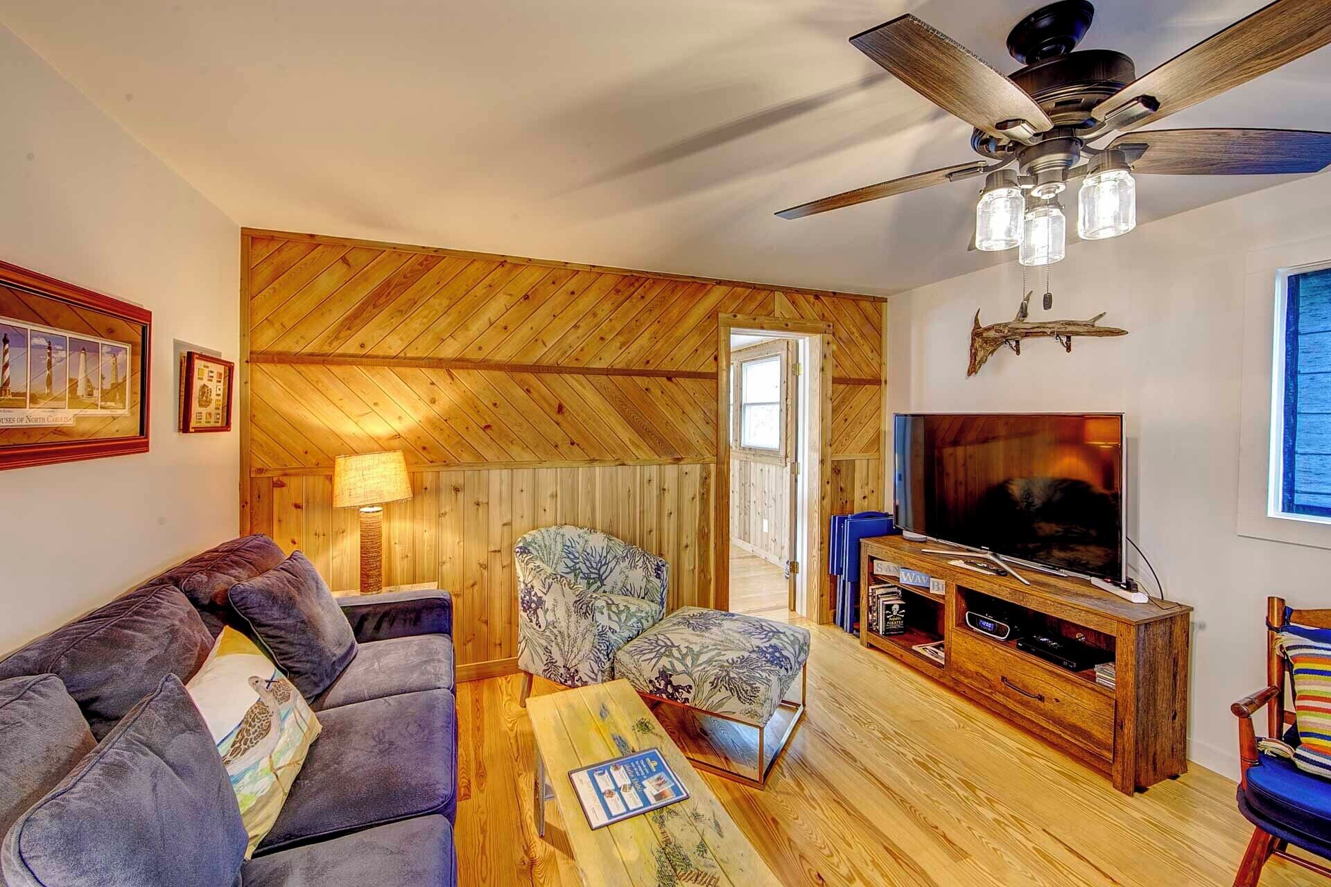 Un salon chaleureux avec un plafond en bois, un ventilateur de plafond, un téléviseur, un canapé, un fauteuil et un meuble de rangement en bois.