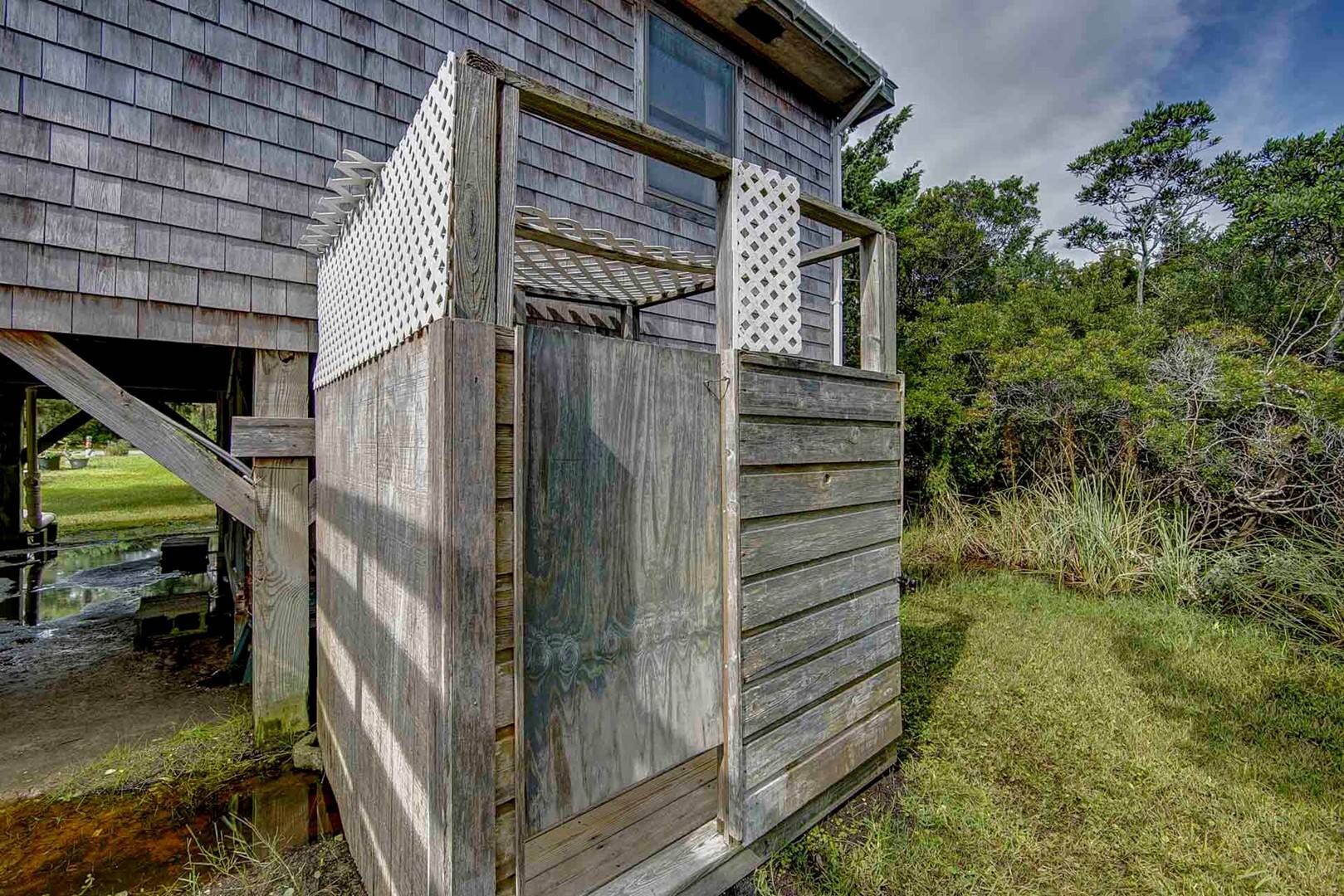 A rustic wooden structure with a lattice window and a door, surrounded by natural vegetation.