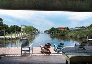 A wooden dock with three Adirondack chairs facing a calm river, surrounded by greenery and houses.