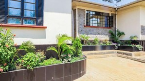 Un pati amb un camí pavimentat, una paret de pedra amb vegetació i un edifici amb una finestra i una porta.