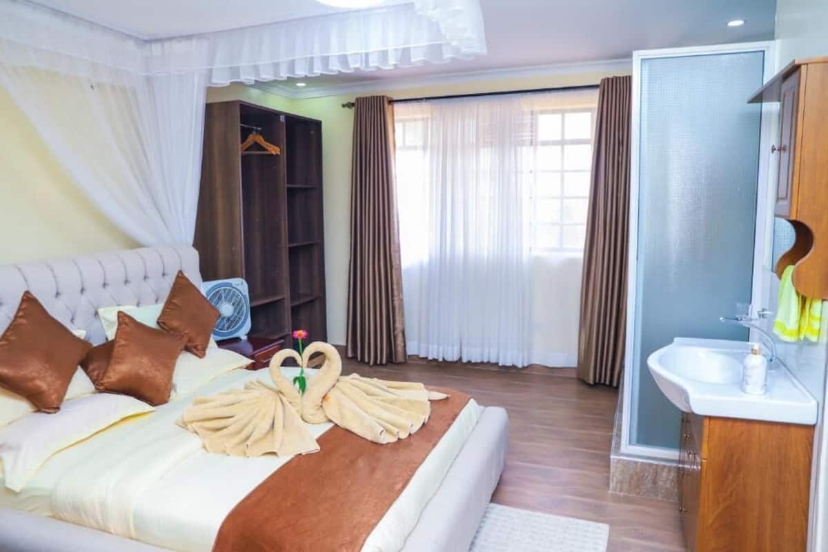 Habitación de hotel con cama, mesita de noche, ventilador, armario, baño con lavabo y espejo, y zona de ducha.