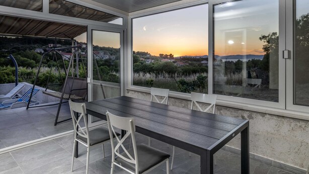 Ein Essbereich mit Tisch und Stühlen, große Fenster mit Blick auf einen Sonnenuntergang und die Landschaft, und eine Schaukelanlage draußen.
