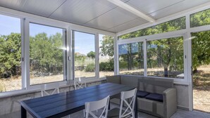 Eine überdachte Terrasse mit einem dunklen Holztisch und weißen Stühlen, ein Korbsessel mit Kissen sowie große Fenster mit Blick auf Grünflächen und Bäume.