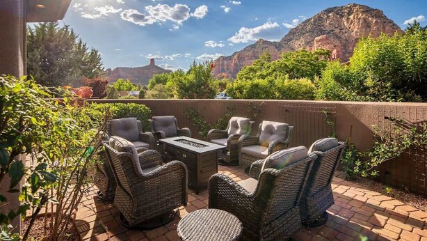 An outdoor patio with wicker furniture and a fire pit, surrounded by greenery and mountains.