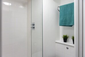 A modern bathroom with white tiles, a glass shower enclosure, a teal towel, and two potted plants on a shelf. - Prince Consort Village (London)