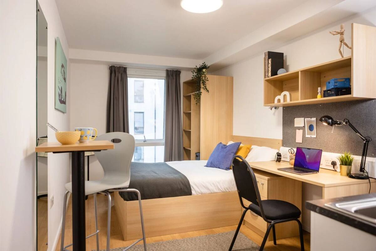 A compact hotel room with a bed, a desk, a chair, a laptop, and a small kitchenette.