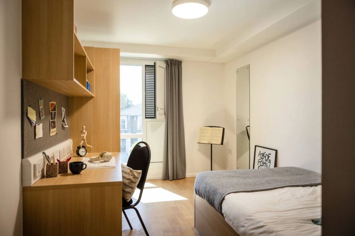 A hotel room with a bed, a desk with a chair, a lamp, and a window with curtains.