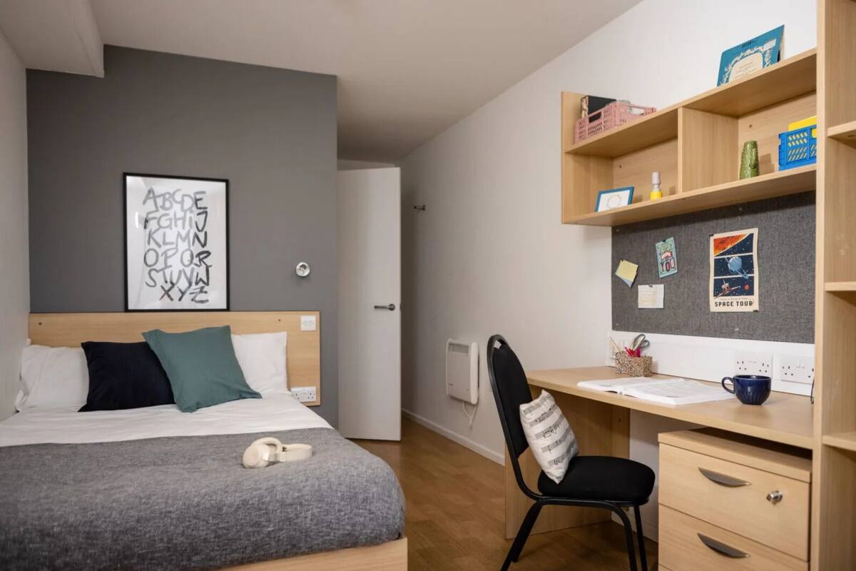 A bedroom with a bed, a desk, and a chair. There is a wall with a poster and a shelf with various items.