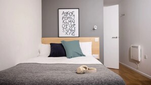 A bedroom with a bed, pillows, a framed alphabet art piece, a door, and a wall-mounted thermostat. - Prince Consort Village (London)
