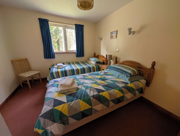 A bedroom with two beds, a chair, a window with curtains, and a lamp on a bedside table. - Pine Croft Cottage - sleeps 4, tucked away in the woodland, 5 minutes from beach (Goonhavern)