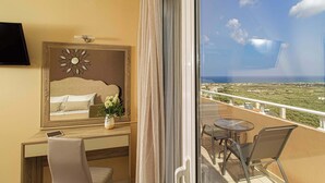 A hotel room with a balcony, a bed with a headboard, a bedside table with a clock, a TV mounted on the wall, and a view of the landscape. - Amoena Villa - Theo Villas With Sea View (Marathokefala)