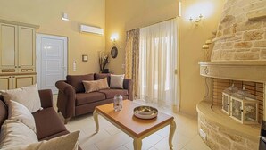 A living room with a sofa set, a coffee table with a decorative bowl, a stone fireplace with birdcages, and a door with a light switch. - Amoena Villa - Theo Villas With Sea View (Marathokefala)