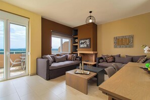A modern living room with a sofa set, a coffee table, and a dining table. There is a fireplace, a balcony with a view, and a large window.