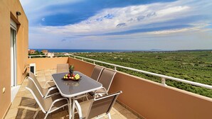 Un balcon avec une table et des chaises, un panier de fruits et une vue sur un paysage verdoyant et la côte.