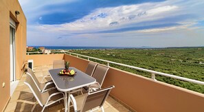 Een balkon met een tafel en stoelen, een fruitmand en uitzicht op een groen landschap en de kustlijn.