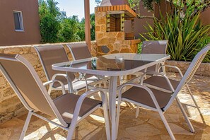An outdoor patio with a stone table and metal chairs, surrounded by a stone wall and greenery.