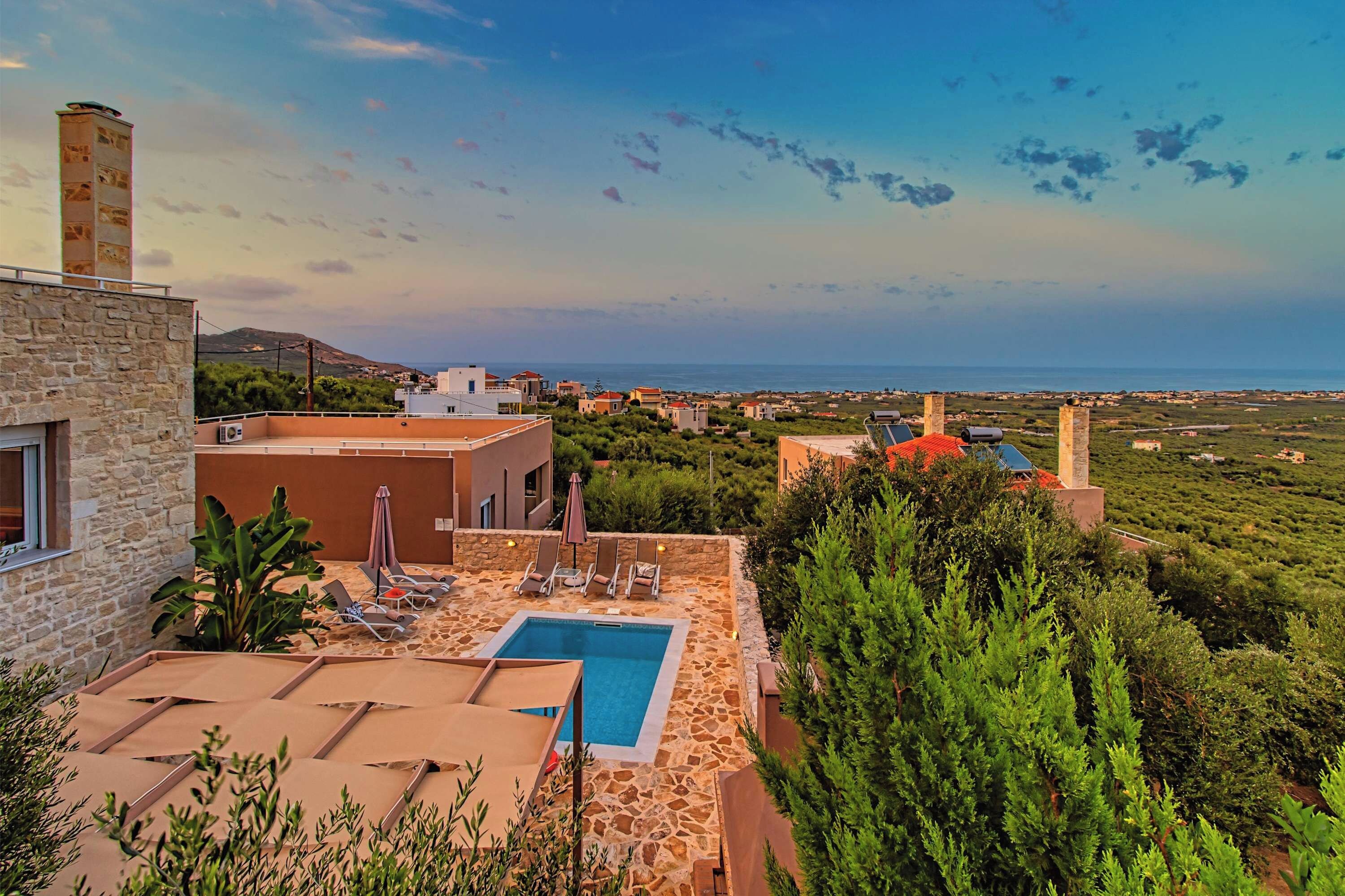 A pool area with lounge chairs and a stone wall, overlooking a coastal landscape with buildings and greenery.