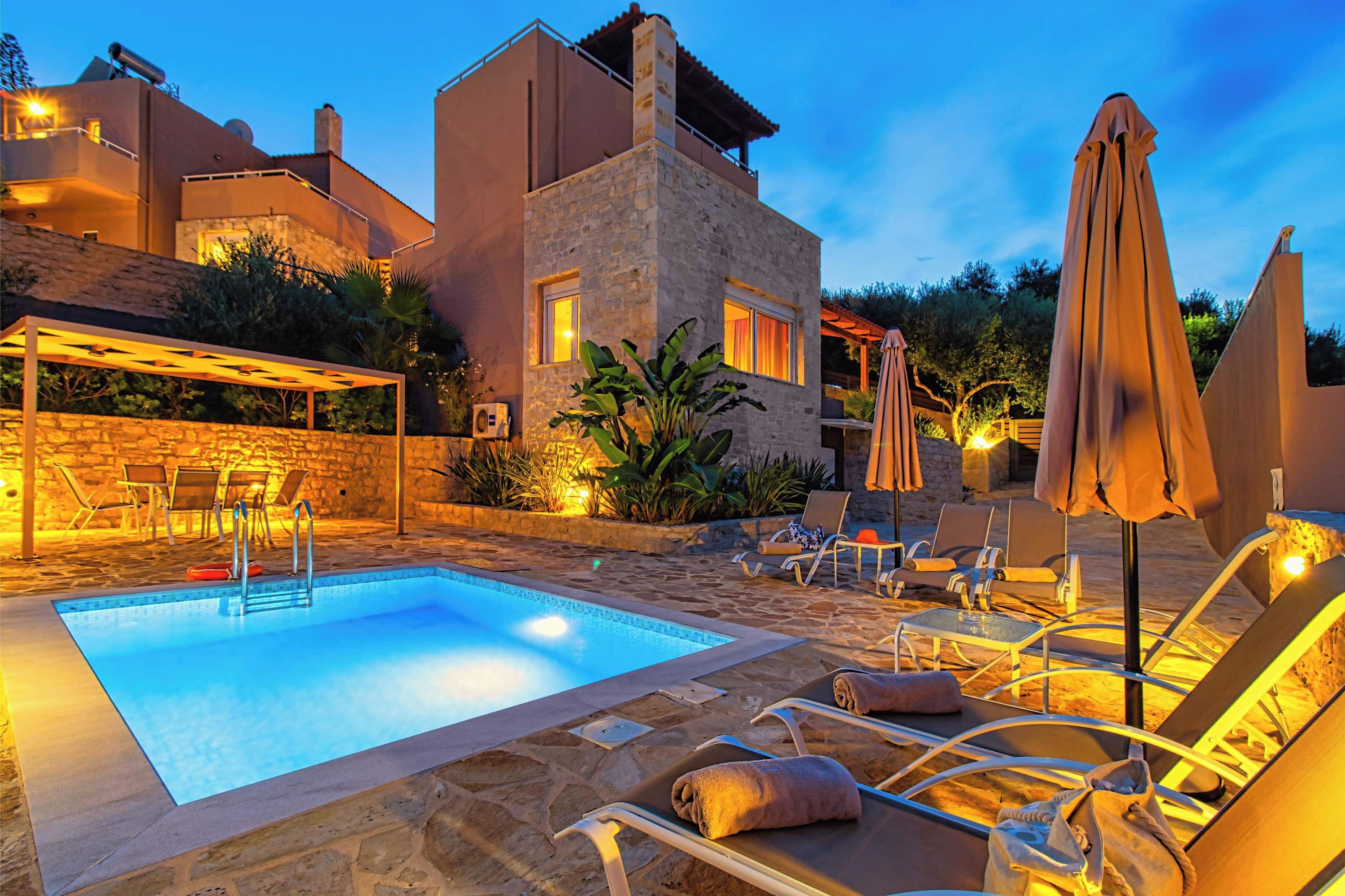A pool area with lounge chairs and umbrellas, stone walls, and a stone building with windows.