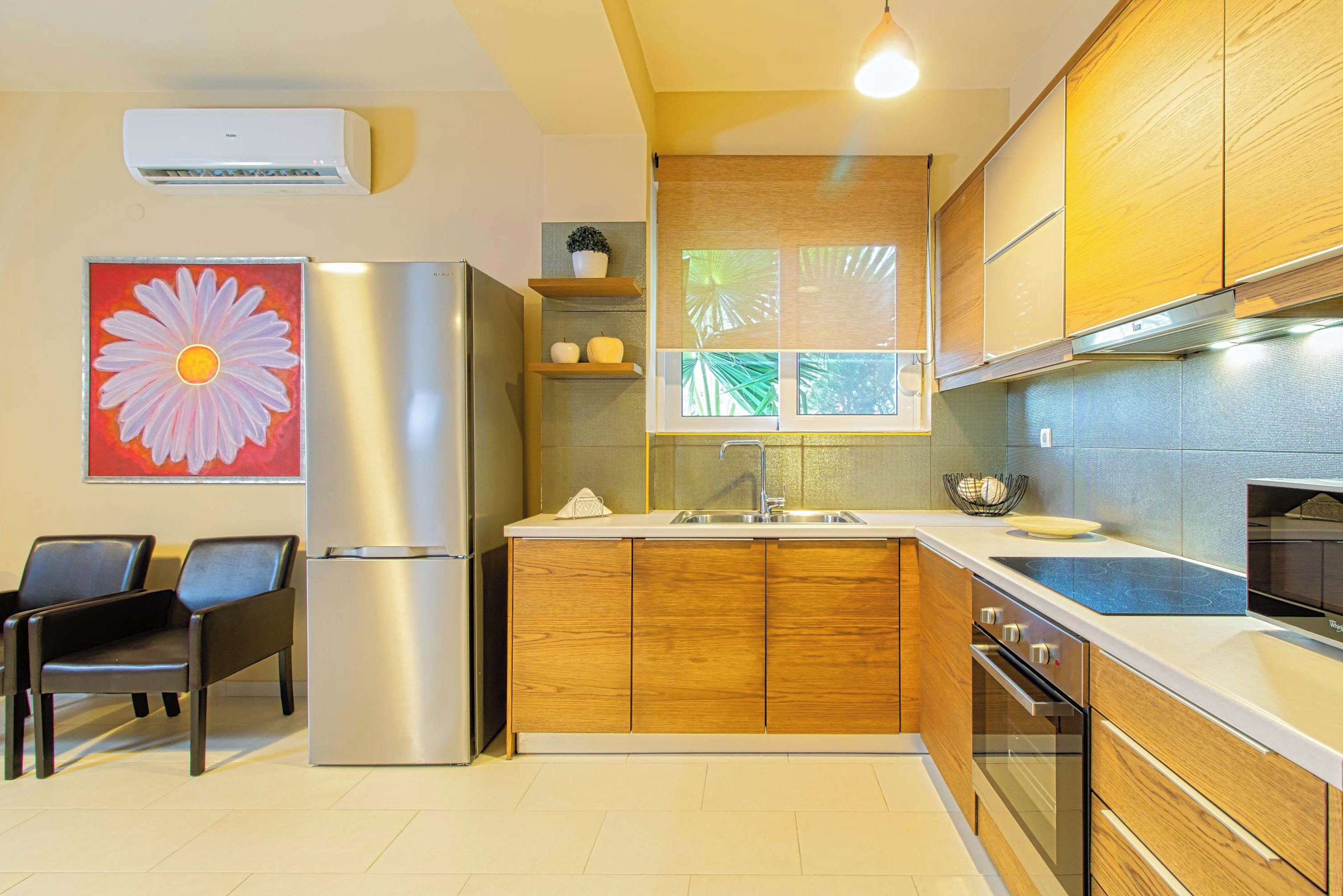A modern kitchen with wooden cabinets, a stainless steel refrigerator, a built-in oven, and a window with a view of palm trees.