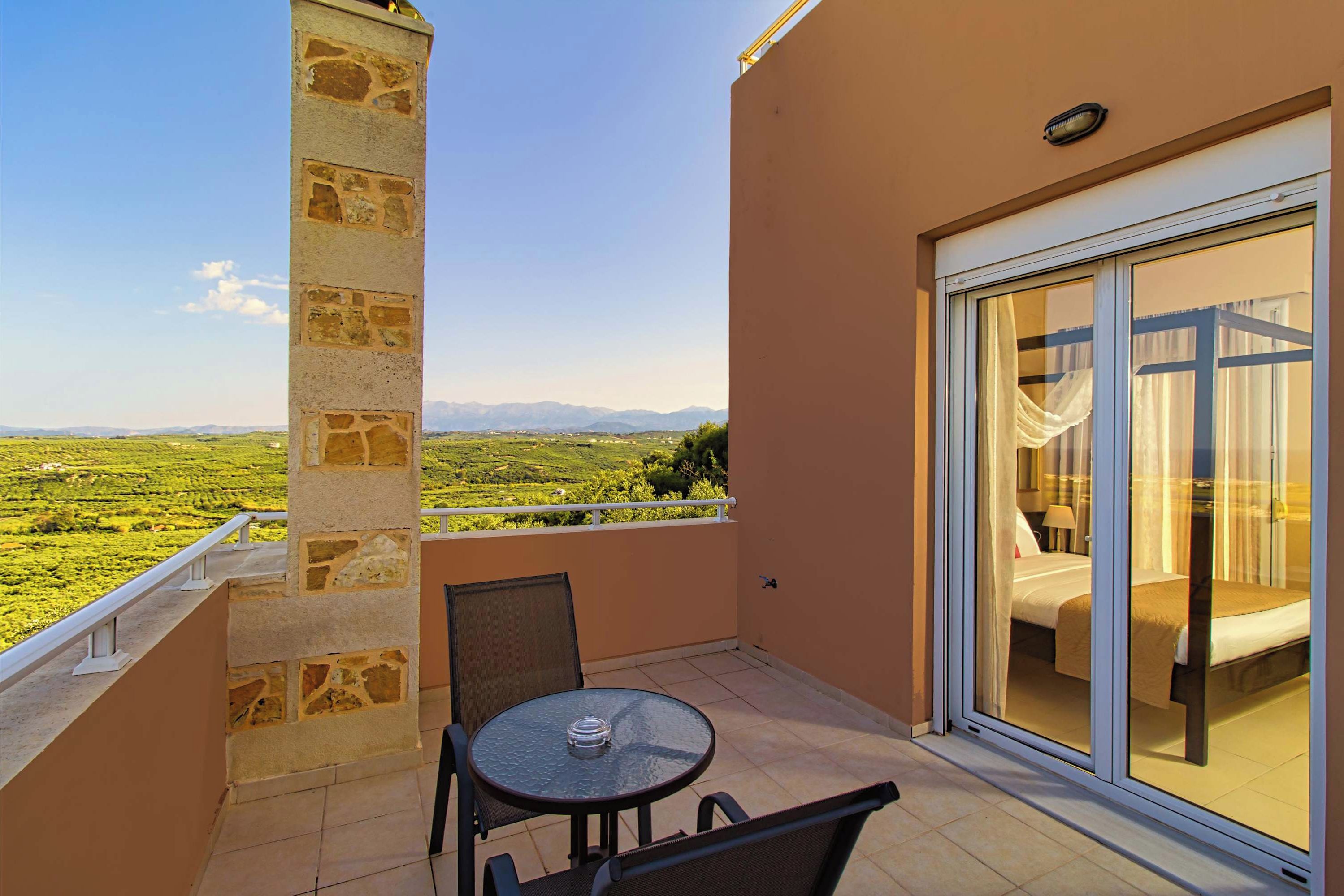 A balcony with a table and chairs, a bed with curtains, and a view of a green landscape with mountains in the distance.