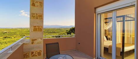 A balcony with a table and chairs, a bed with curtains, and a view of a green landscape with mountains in the distance.