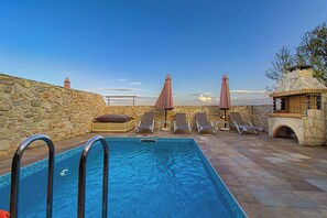A swimming pool with lounge chairs and umbrellas, stone walls, and a stone fireplace.