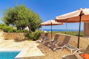 Zona de piscina con sillones de descanso y sombrillas, suelo de piedra y vista al mar.