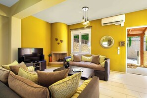 A living room with a sofa set, a flat-screen TV, a fireplace, and a large window with a view of greenery. - Lillium Villa - Theo Villas With Sea View (Marathokefala)