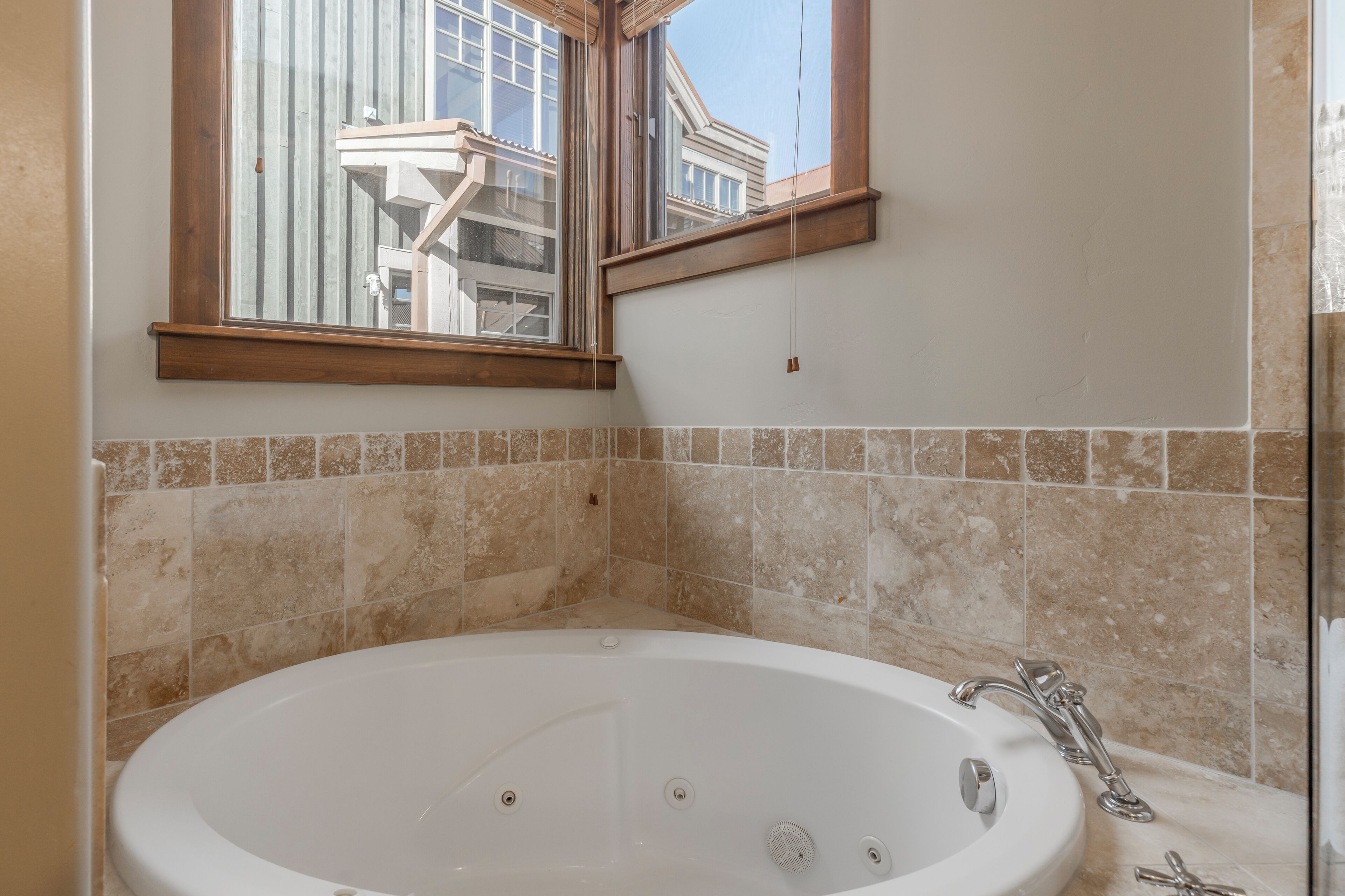 A bathroom with a large bathtub, a window, and beige tiled walls.