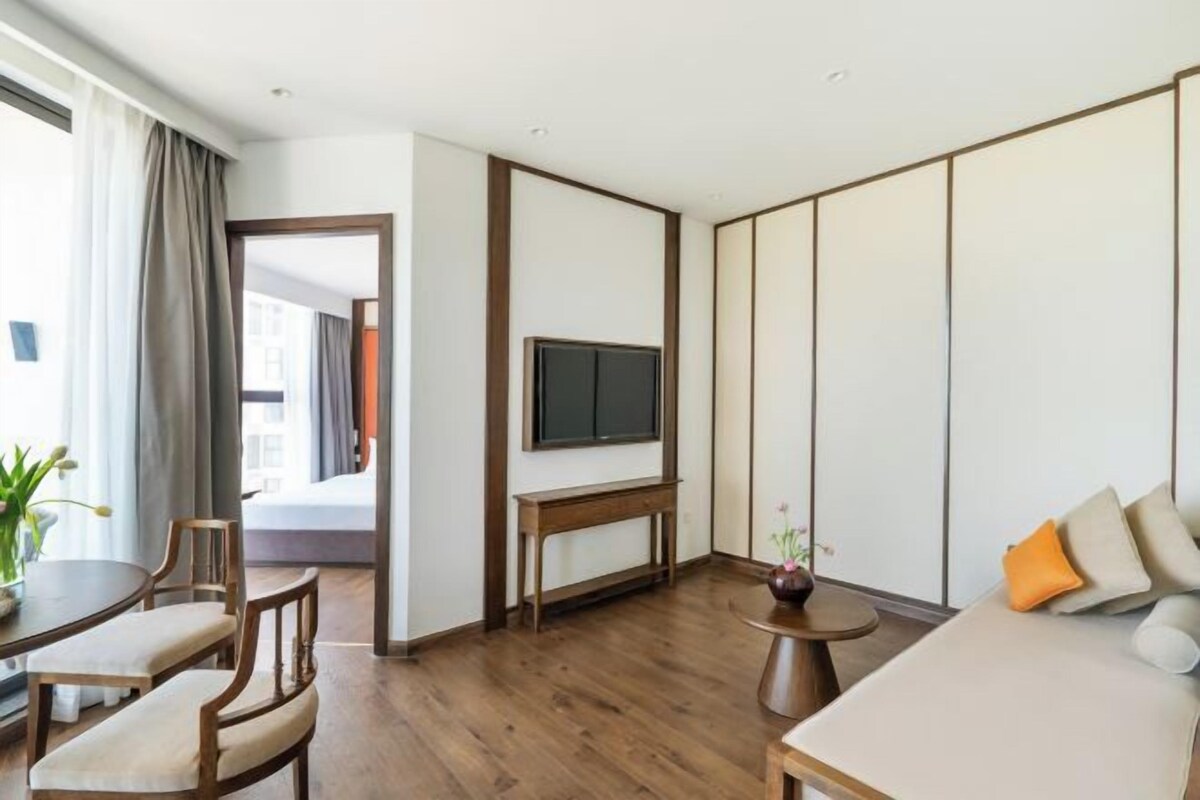 A modern hotel room with a wooden floor, a sofa, a dining table with chairs, a TV mounted on the wall, and a view of the outside.