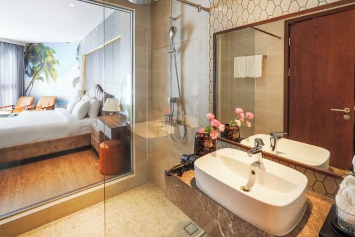 A modern hotel room with a large bed, a glass shower enclosure, a vanity with two sinks, and a large mirror.