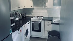 Pieni, käytännöllinen keittiö, jossa on valkoiset kaapit, kaasuliesi, pesukone ja roskakori.