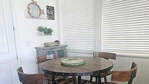 Een eetgedeelte met een ronde houten tafel, vier stoelen, een decoratief wandobject en een raam met jaloezieën.