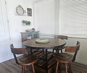 Un comedor con una mesa redonda de madera, cuatro sillas, un adorno en la pared y una ventana con persianas.