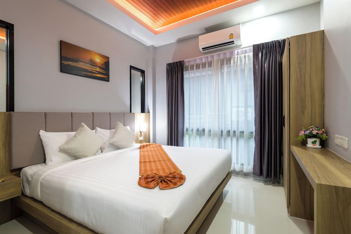 A hotel room with a bed, a nightstand, a wooden headboard, a wall-mounted air conditioner, a window with curtains, and a small plant on a wooden shelf.